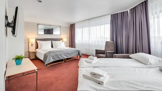 A hotel room with two neatly made beds, modern furniture, and large windows draped with sheer and opaque curtains.