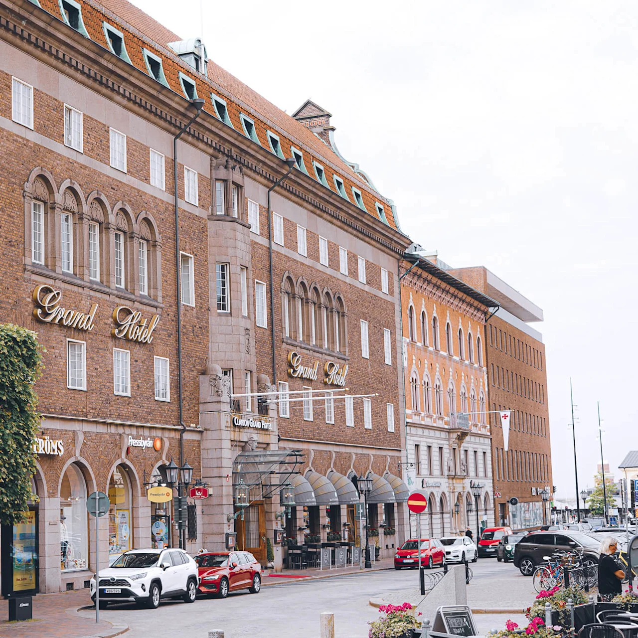 Et stort mursteinshotell, "Grand Hotel," med folk og biler på en bygate.