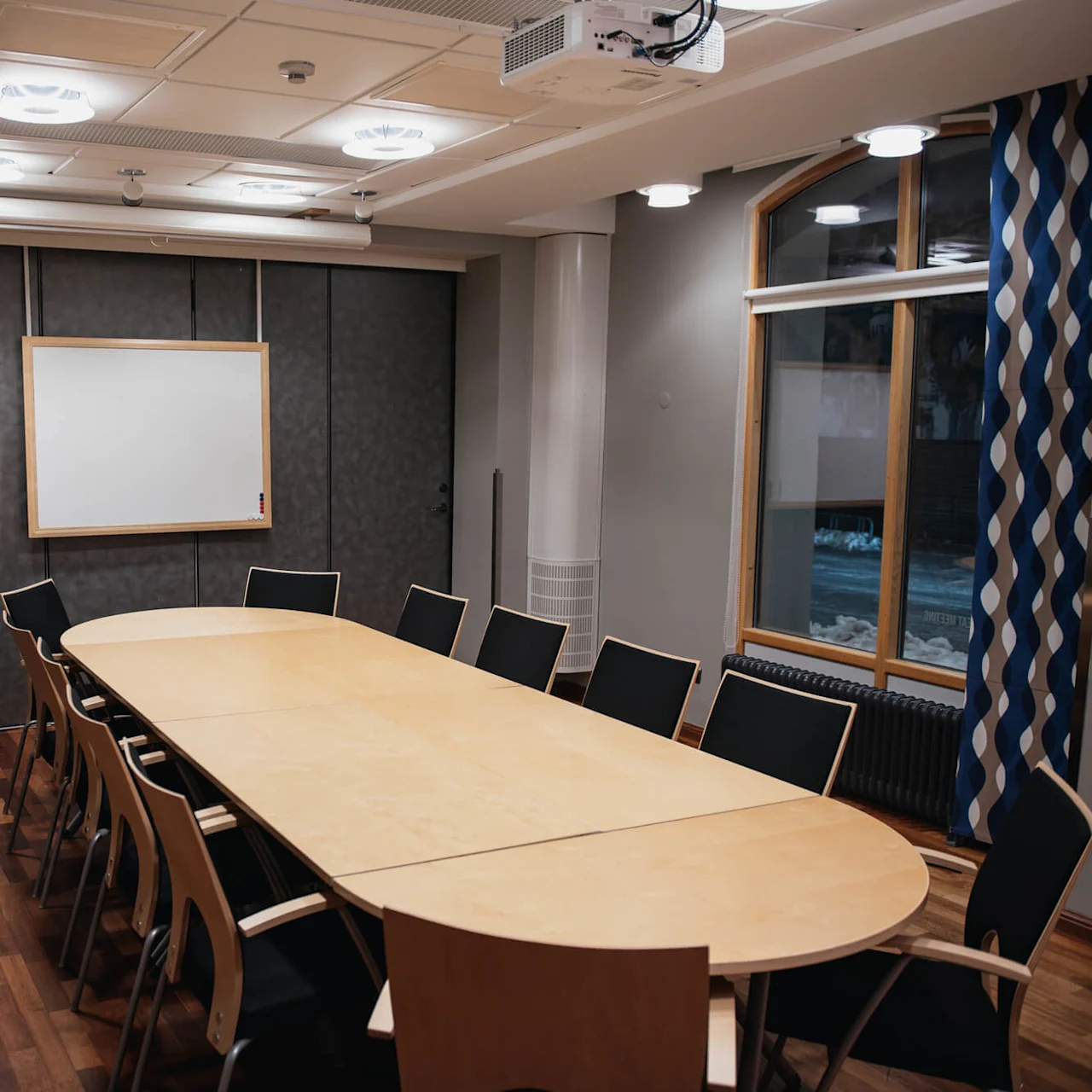 A conference table with chairs is set in a room with wooden flooring, a whiteboard on the wall, overhead lights, a window with blue curtains, and a ceiling-mounted projector.
