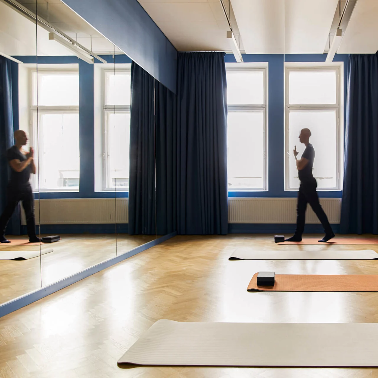 Yogamåtter ligger på et trægulv i et spejlværelse; en person går ved siden af måtterne. Store vinduer med blå gardiner omkranser baggrunden.