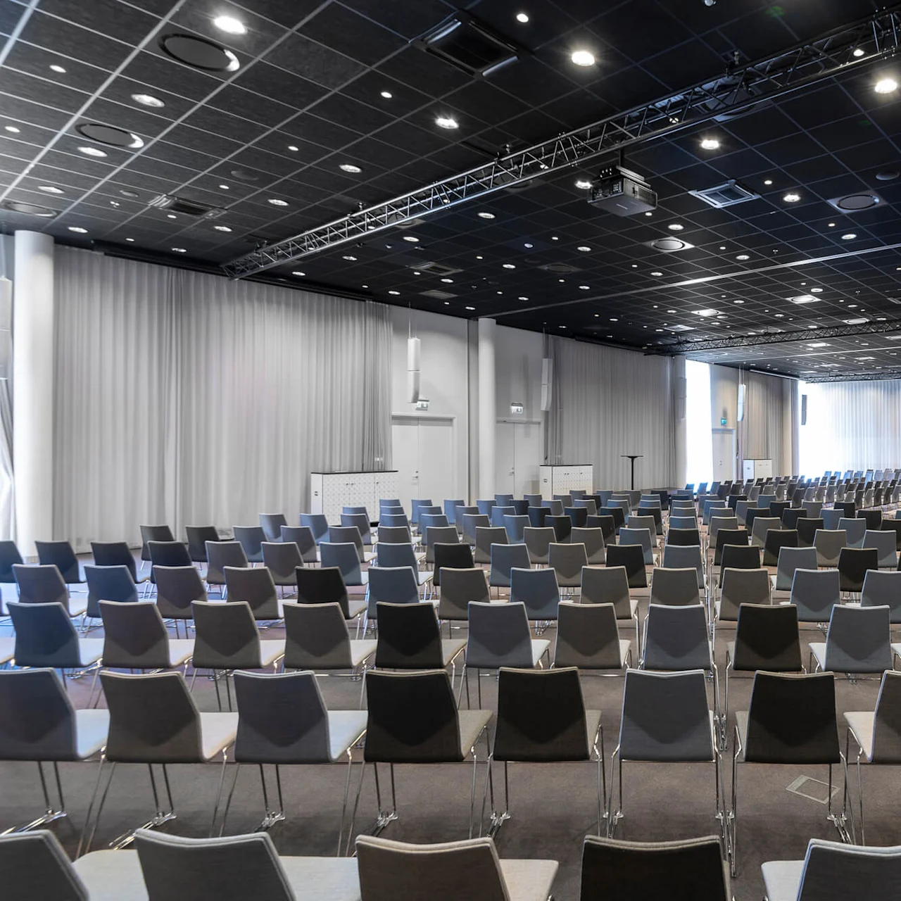 Rows of empty chairs face a stage in a spacious conference room, with white walls and curtains, under a ceiling dotted with recessed lights.