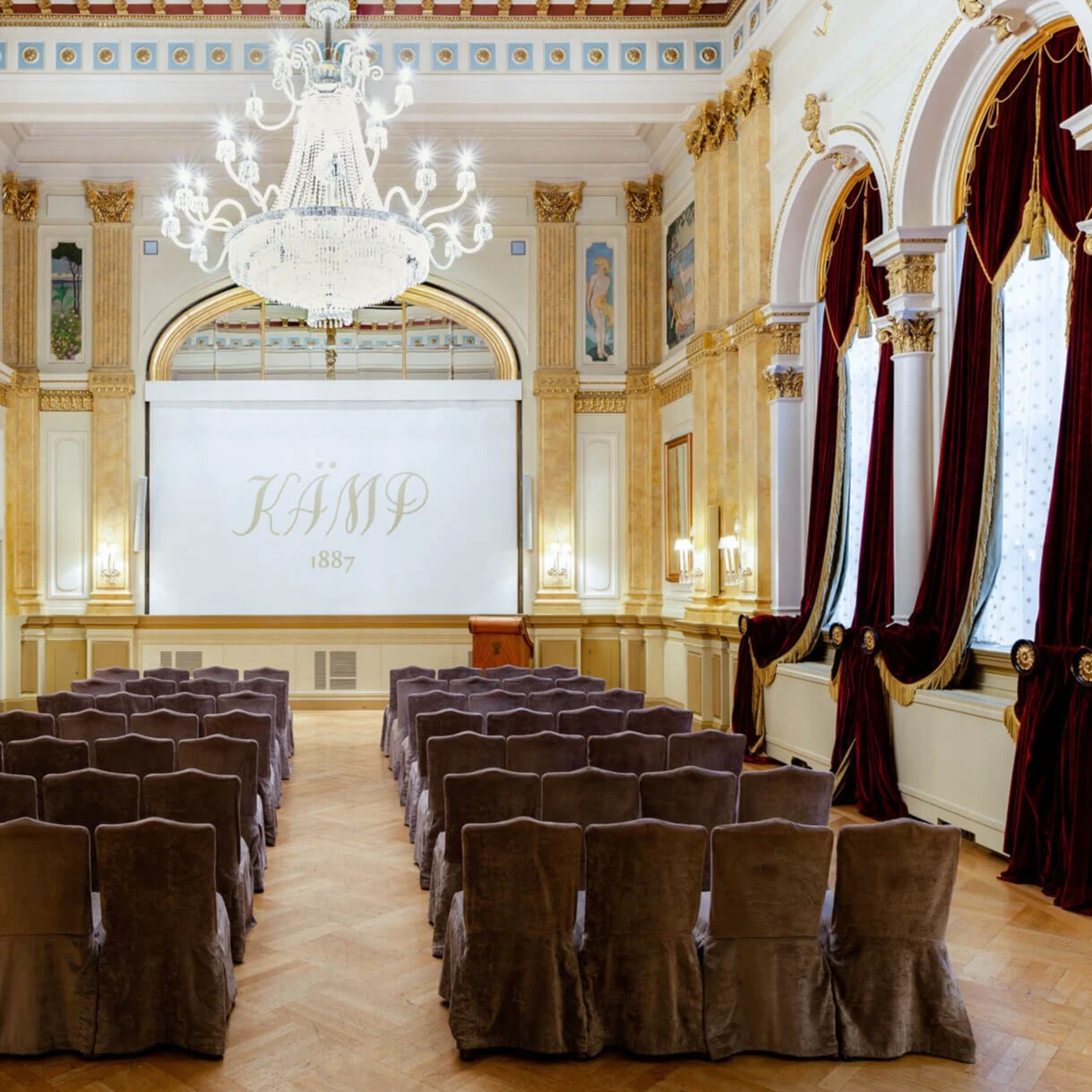 A grand, ornate hall features rows of covered chairs facing a large screen displaying "KÄMP 1887." Chandeliers and large windows with red curtains adorn the elegant space.