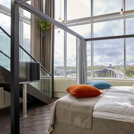 A modern bed with colorful pillows sits under a large window, offering a cityscape view. A TV is mounted nearby. Tall curtains frame the loft-style room.