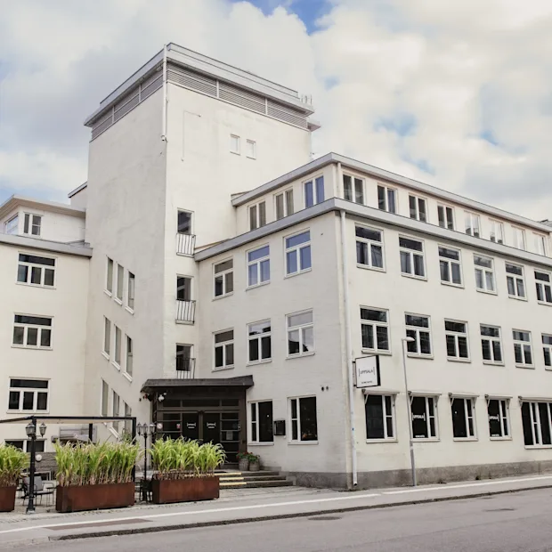 Exterior of a white hotel building with 'home hotel' sign, green plants, and a street in front. Home Hotel Uppsala.
