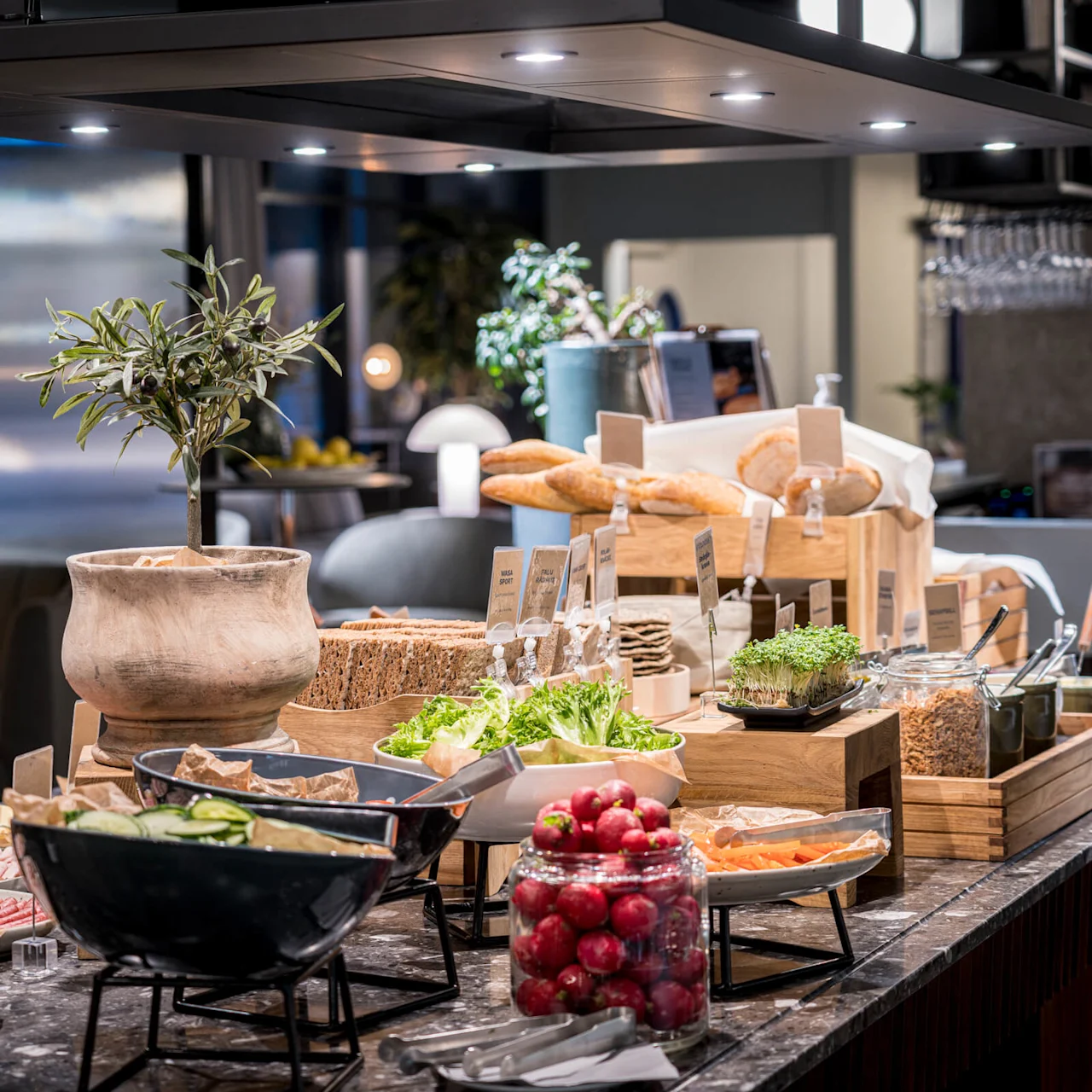 A buffet table displays fresh vegetables, assorted bread, and toppings in a modern restaurant setting, illuminated by spotlights.