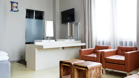 A modern living space featuring two brown leather chairs, a wooden side table, a wall-mounted TV, and a large beige curtain filtering sunlight. A blue letter ‘E’ decorates the wall.