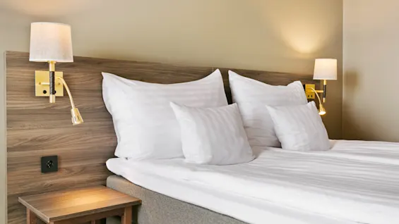 Viihtyisä hotellisänky, jossa on raikkaat valkoiset vuodevaatteet, tyynyjä ja lämmin valaistus seinävalaisimista puisen sängynpäädyn yläpuolella.