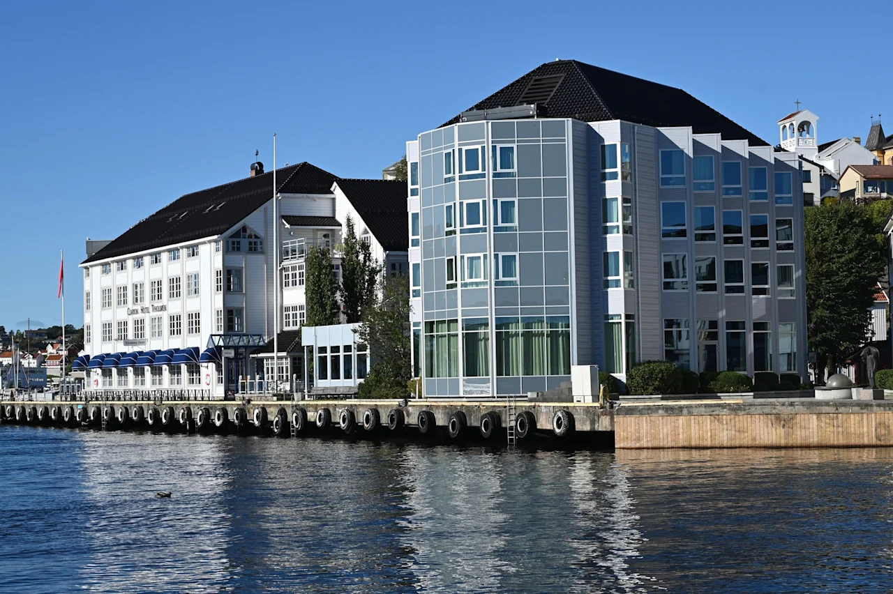 Et hotell med blå markiser og en moderne glassbygning ved vannkanten på en solrik dag. Teksten lyder Hotel.