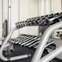 Dumbbells rest on a metallic rack in a gym, reflecting light from nearby mirrors, with exercise equipment surrounding the area.