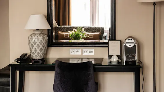 Et skrivebord med en svart stol og en lampe til venstre; en kaffemaskin og innrammet bilde til høyre. Stor speil reflekterer en sofa og bukett, som skaper en koselig atmosfære.