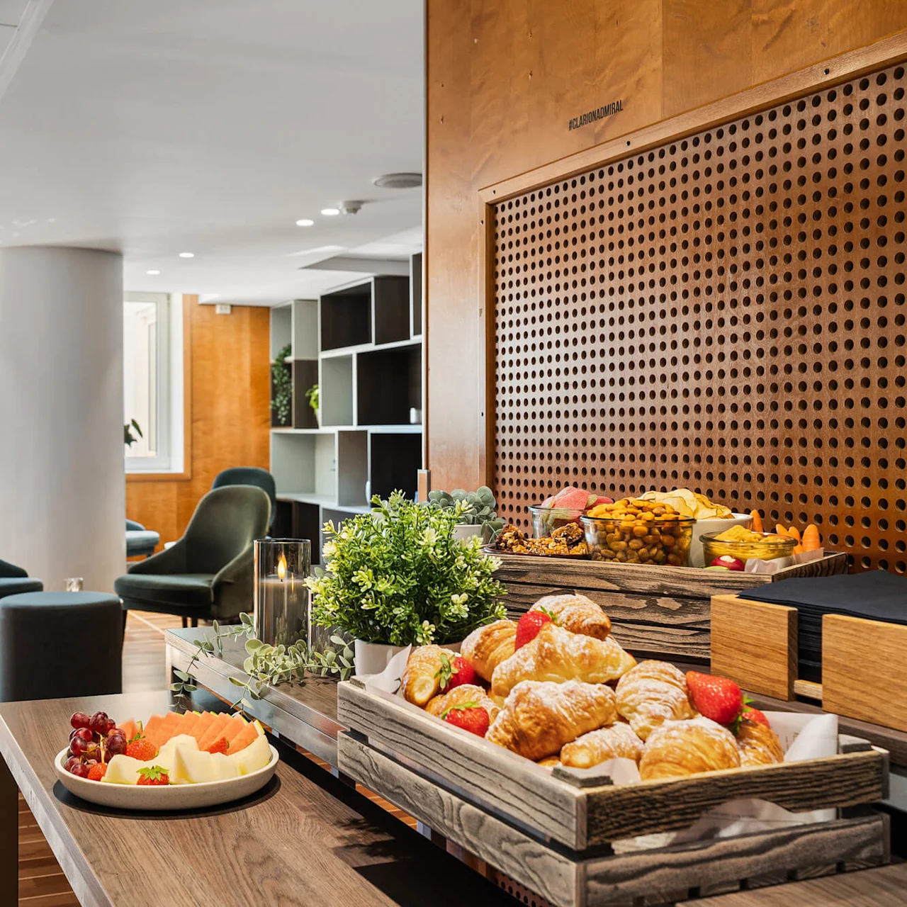 Croissanter og frugt på et træbord i en moderne, lyst oplyst lounge med grønne stole, trævægge og store vinduer. Tekst: "FLAURAUD" ses delvist på en panel.