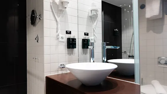 White bowl sink on a wooden countertop with wall-mounted hairdryers and soap dispensers, set against tiled walls in a modern bathroom.