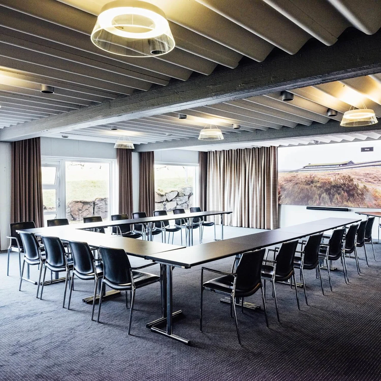 Konferencerum med U-formet bordopstilling og sorte stole; hængende lamper og stort landskabsfoto på væggene; vinduer og gardiner omgiver en udsigt udenfor.