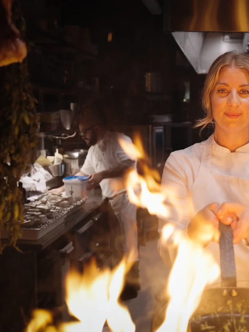 En kock slänger skickligt mat i en brinnande panna i ett livligt kök, med en kollega som arbetar i bakgrunden, omgiven av hängande örter och kött.