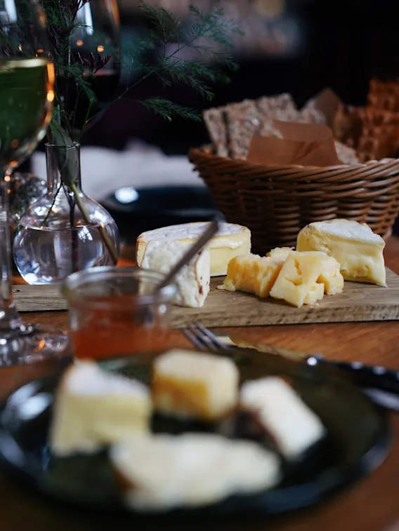 Ostsortiment vilar på en träbräda med brödkorg och vin i närheten; placerat på ett middagsbord med en vas med lövverk i en svagt upplyst miljö.