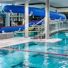 En blå vattenrutschbana slingrar sig ovanför en inomhuspool, omgiven av modern arkitektur med en böjd bro över vattnet. En spiraltrappa syns till vänster.