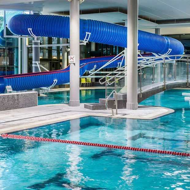 En blå vattenrutschbana slingrar sig ovanför en inomhuspool, omgiven av modern arkitektur med en böjd bro över vattnet. En spiraltrappa syns till vänster.