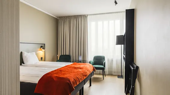 Et koselig hotellrom med en seng pyntet med et oransje pledd. Myk belysning og moderne innredning gir en innbydende atmosfære, med sitteplasser ved vinduet og funksjonelle fasiliteter.