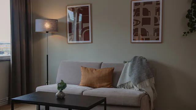 Soffa med kuddar står i ett mysigt vardagsrum, flankerat av ett svart soffbord och upplyst av en golvlampa. Två abstrakta konstverk hänger på den beige väggen.