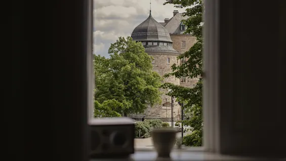 Ett slott syns genom ett fönster, med en kaffekopp och en bok på fönsterbrädan. Bakgrunden har lövträd och en delvis molnig himmel.