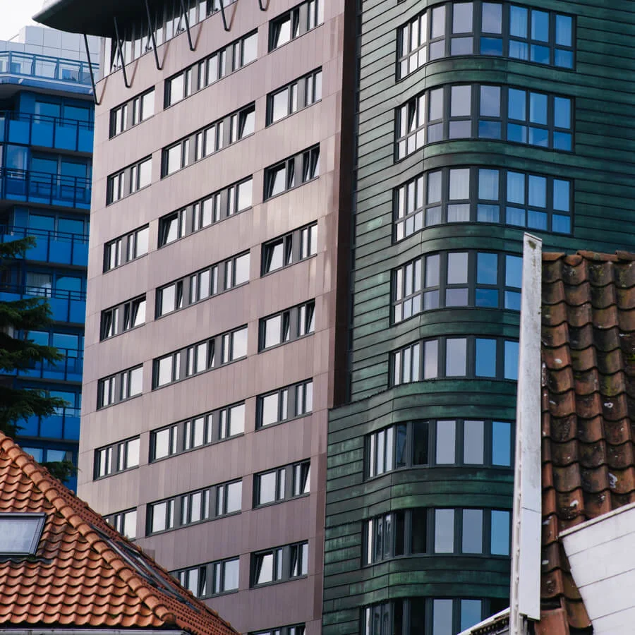 Tall building with modern architecture featuring curved and flat sections, multiple windows, and contrasting colored materials. Surrounded by traditional rooftops and other urban structures.