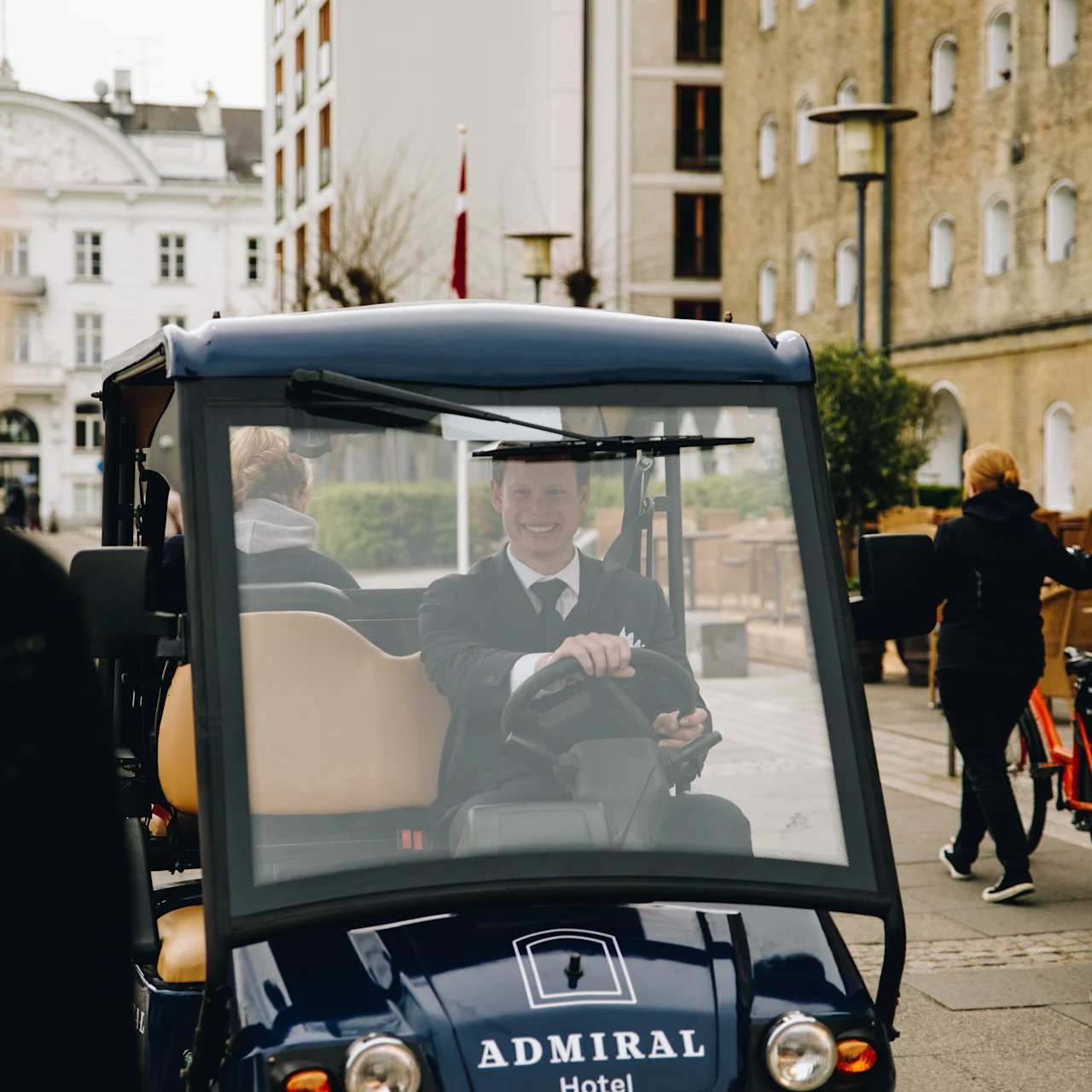 A person in a suit drives a small vehicle with "ADMIRAL Hotel" on it through a busy street, where people walk and cycle near brick and white buildings.