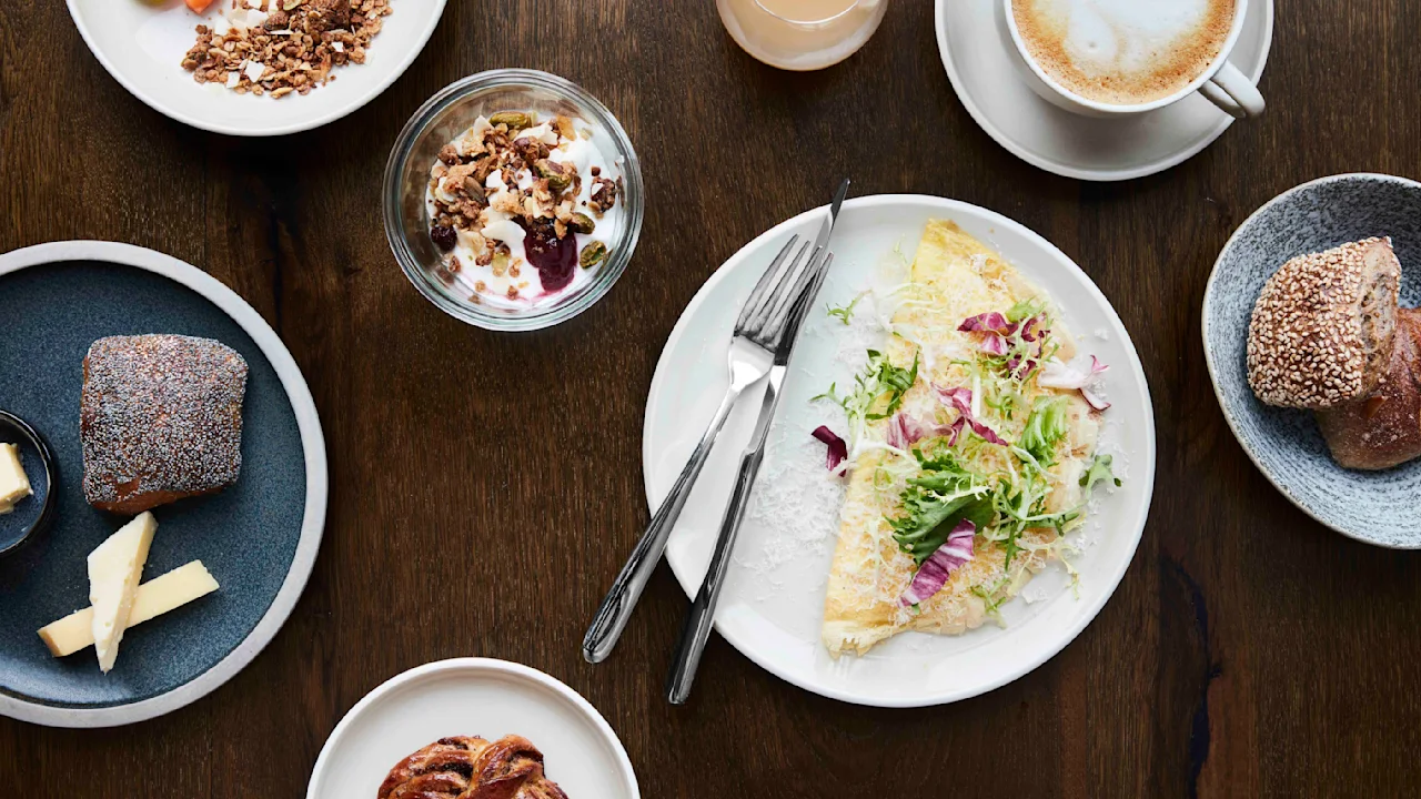 Omelett på en hvit tallerken garnert med grønt og rødløk, omgitt av en kaffekopp, juice, yoghurt med granola, bakverk, ost og frukt på et trebord.