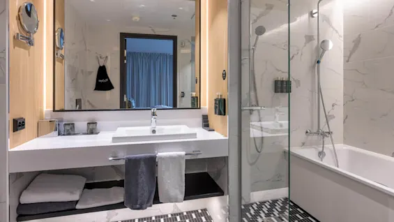 A modern bathroom features a white sink with towels below, a mirrored wall, and a glass-enclosed shower-bathtub combo. Marble tiles and contemporary fixtures create a sleek ambiance.