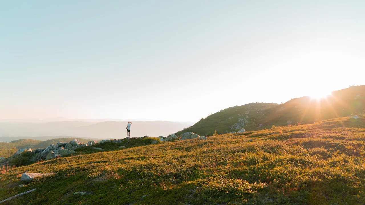 En person står på en gresskledd bakketopp under soloppgang, omgitt av fjerne fjell og en klar himmel.