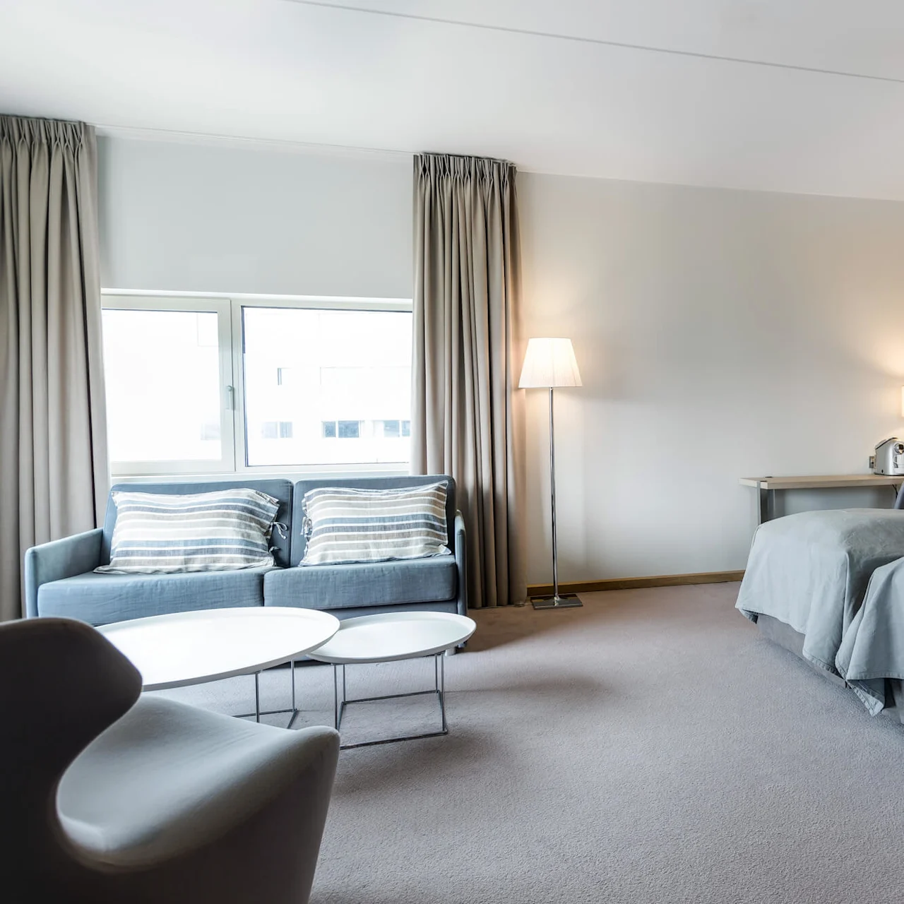 A hotel room features two neatly made twin beds, a gray sofa with striped pillows, and a white coffee table. Floor lamps and a desk with a coffeemaker complete the modern decor.