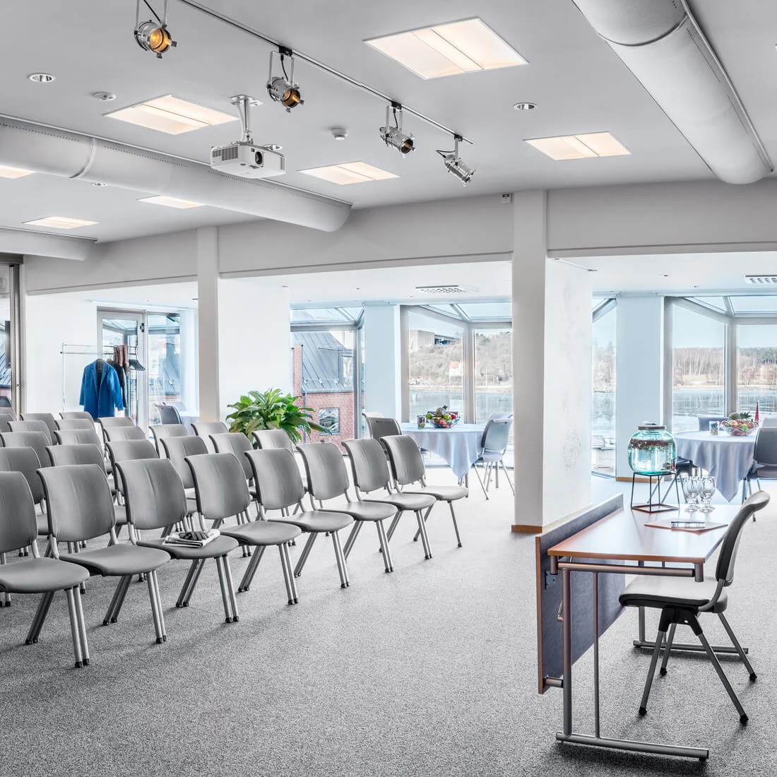 Rows of gray chairs are arranged in a conference room with red curtains. Tables with refreshments are near large windows overlooking a waterfront. Potted plants decorate the bright space.