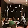 People sit at round tables, engaged in conversation and dining in a warmly-lit restaurant with hanging globe lights and red carpeting.