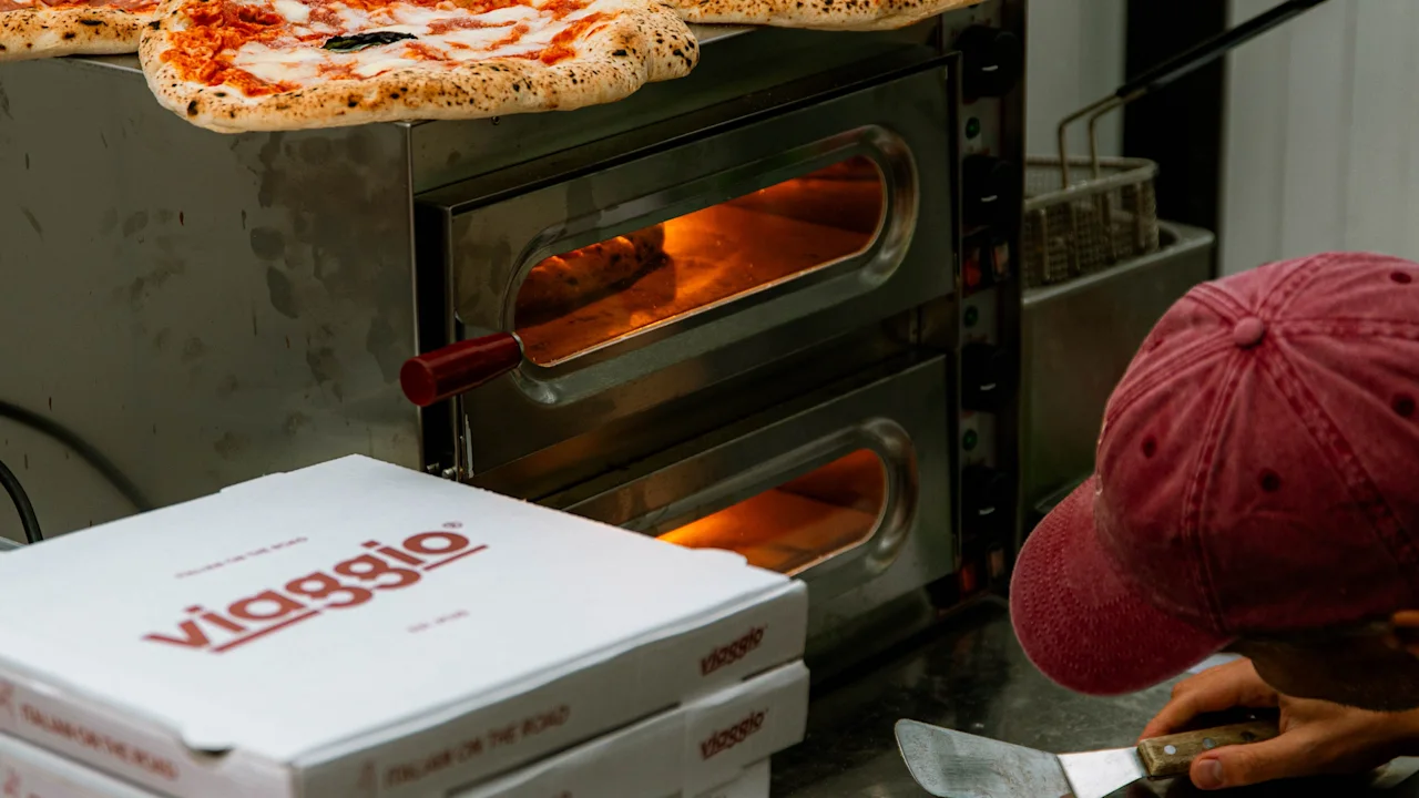 Pizzor ligger på en ugn medan en person kontrollerar dem, omgivna av staplade "viaggio" pizzakartonger utomhus. Text på kartongerna: "ITALIAN ON THE ROAD."