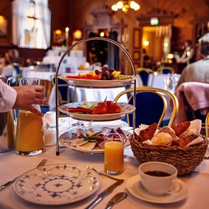 Tiered stand displays fruit, smoked salmon, and cheese; a hand pours orange juice. Elegant dining room with chandeliers and blue chairs in the background.