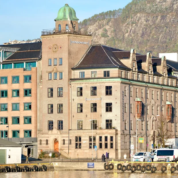 A charming waterfront scene featuring a historic building with a green dome and the text HAVNEKONTORET, against a mountain backdrop.