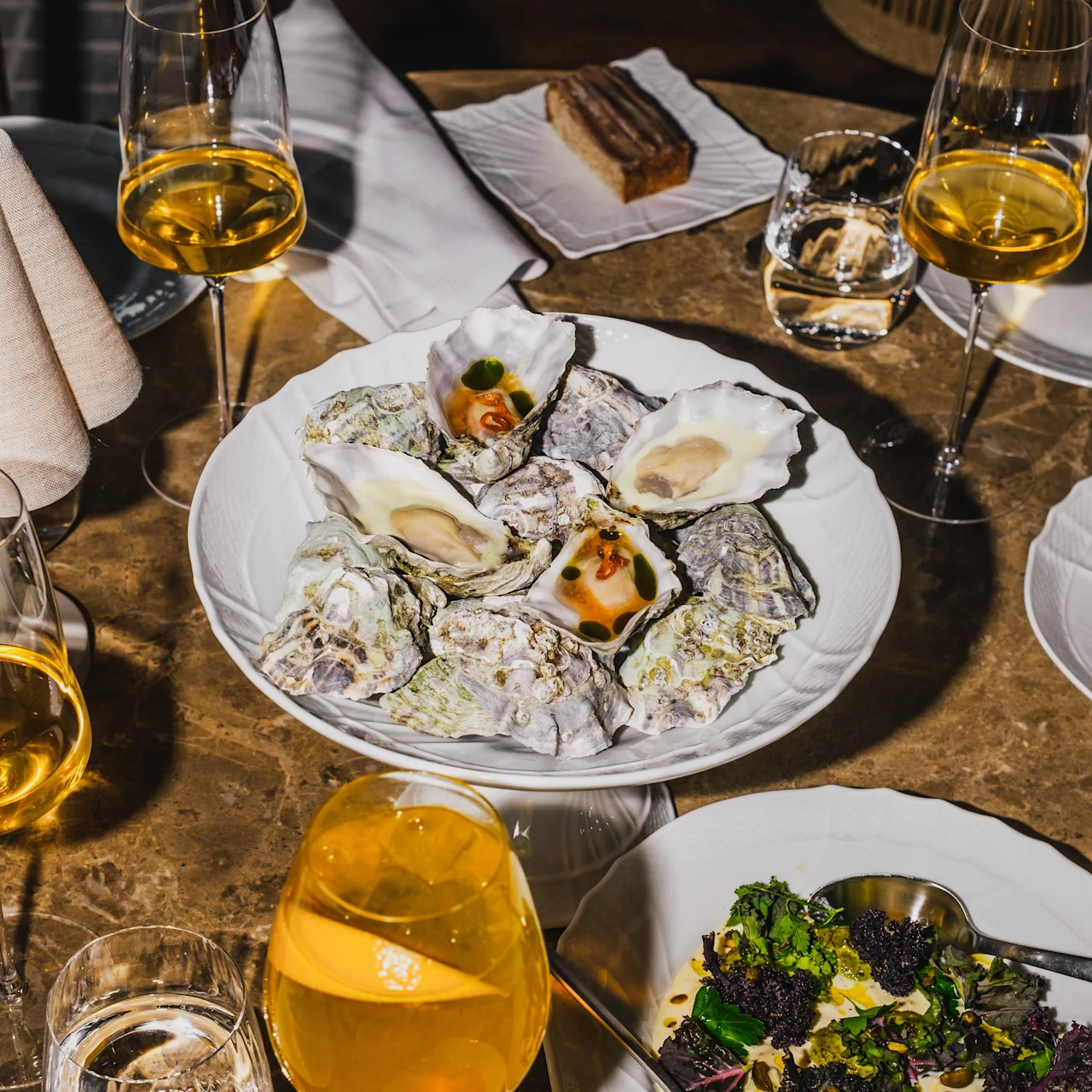 Oysters arranged on a decorative plate; surrounded by wine glasses, a slice of pastry, and a bowl of salad; placed on a wooden table in a restaurant setting.