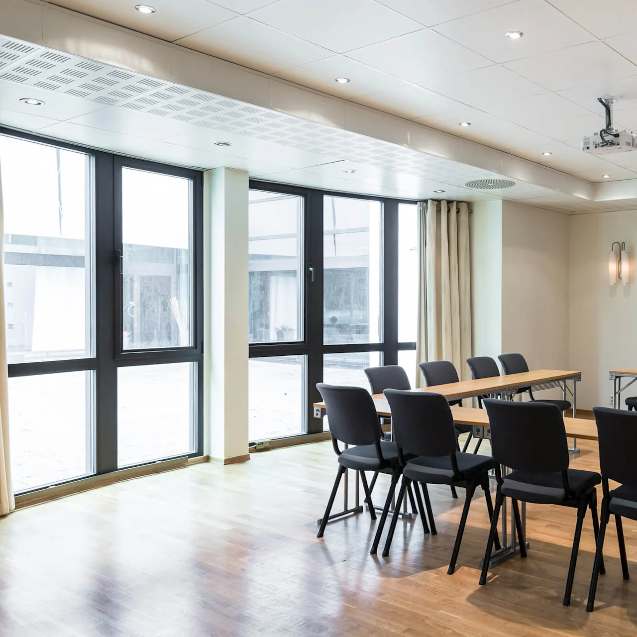 Conference room featuring empty chairs around tables, with a large screen on the wall; brightened by natural light from large windows.
