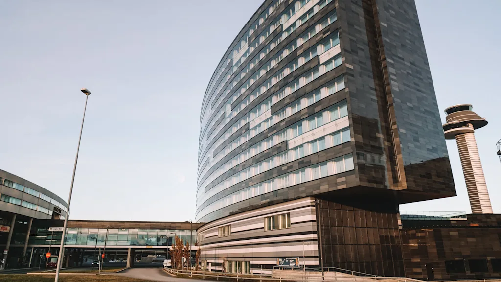 Ett högt, modernt hotell med glasfasad står bredvid en flygplatskontrolltorn under en klar himmel. En gångbro förbinder det med intilliggande byggnader.