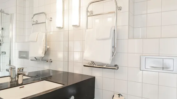 Ett rent, modernt badrum med vita kaklade väggar, en svart kommod och fräscha vita handdukar på handdukstorkar.