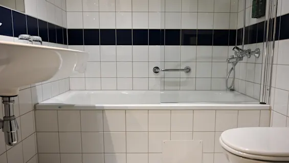 Badkar med en glasavskärmning och silverhandtag, intill en vit kakelvägg med mörkblå rand; i närheten, ett handfat och toalett kompletterar badrumsmiljön.