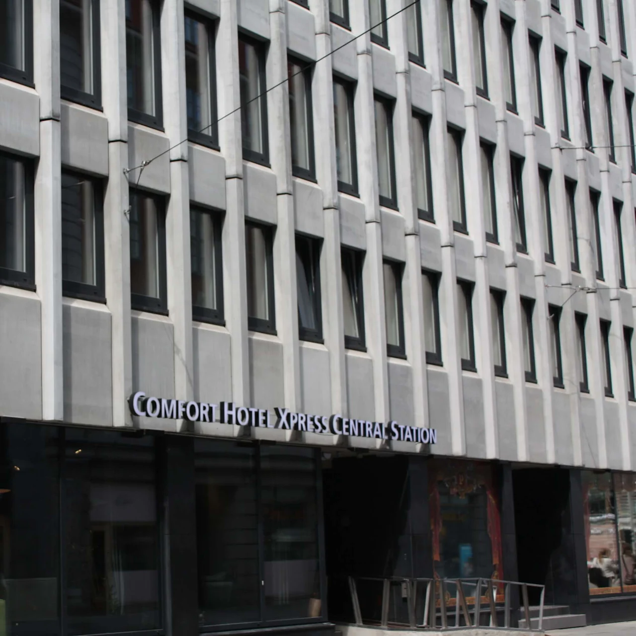 A modern, gray building facade with multiple windows hosts a sign reading “Comfort Hotel Xpress Central Station.” The urban setting includes large ground-floor windows and a minimalistic design.