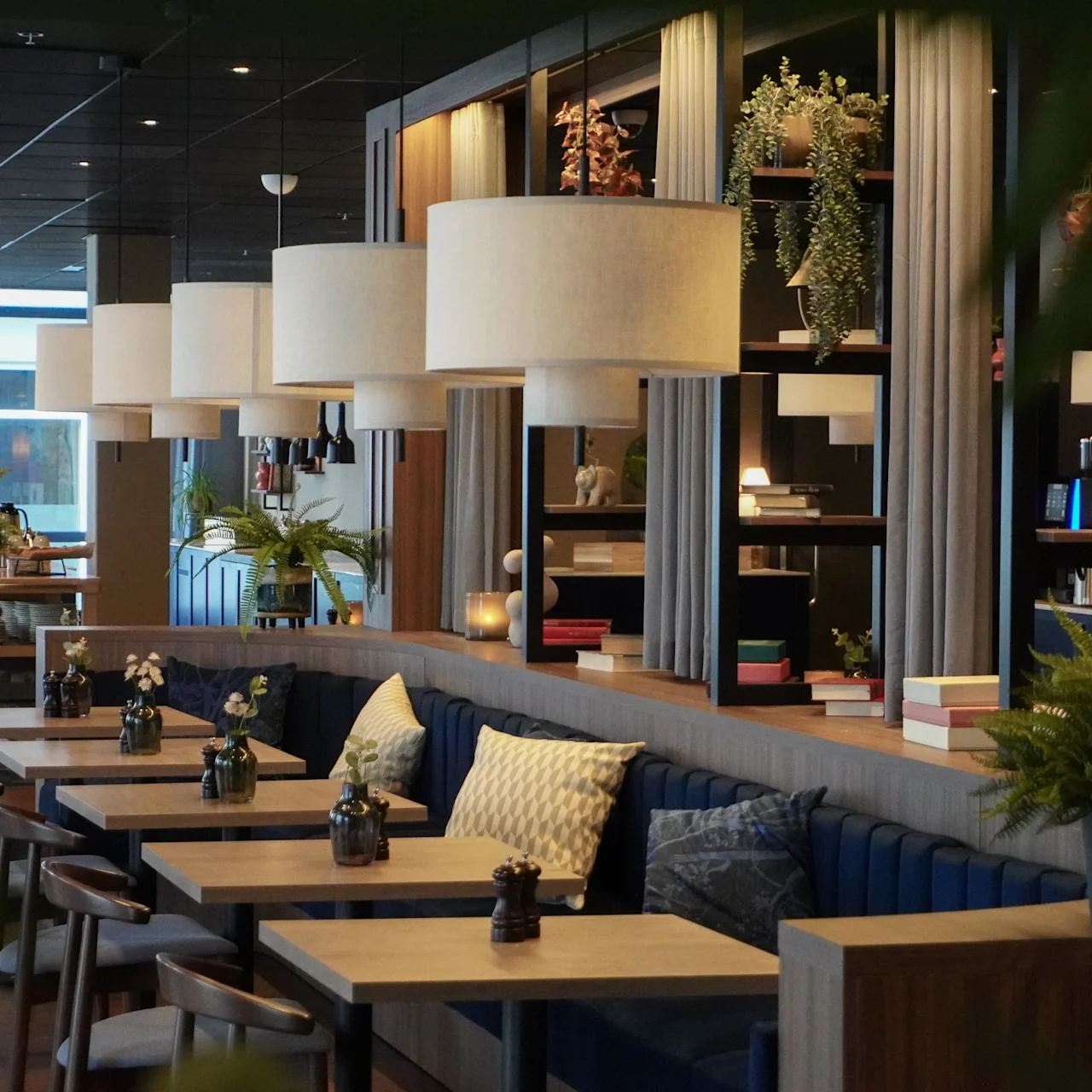 Bord kantar ett stilrent, modernt café; hängande lampor belyser krukväxter och böcker på trähyllor, vilket skapar en mysig atmosfär med vadderade sittplatser.