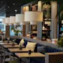 Bord kantar ett stilrent, modernt café; hängande lampor belyser krukväxter och böcker på trähyllor, vilket skapar en mysig atmosfär med vadderade sittplatser.