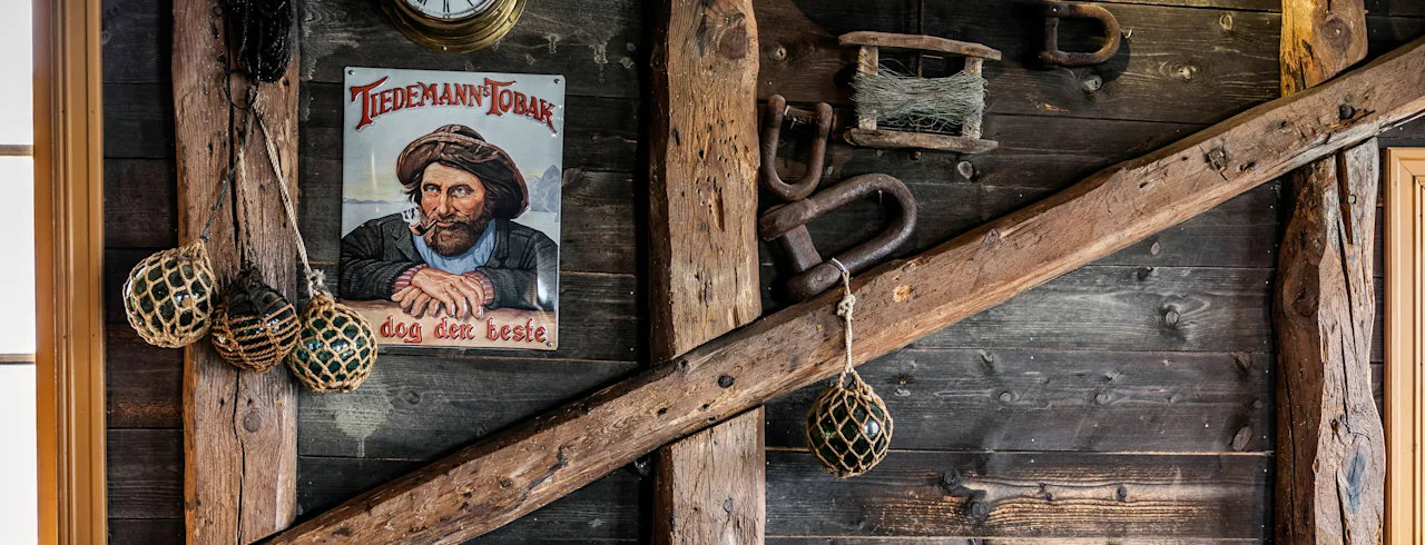Ruokapöytä, jossa laseja ja lautasia; nahkatuolit seisovat rustiikkista puuseinää vasten, koristeltu vanhanaikaisella "Tiedemann Tobak god det beste" julisteella ja kalastuskoristeilla.