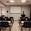 A modern conference room with rows of tables and chairs facing a large projector screen, ready for a presentation.