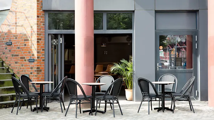 Outdoor café tables with black chairs are arranged on a cobblestone surface. A red brick wall and a large pillar form the background, with a doorway leading inside.