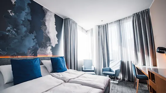 A hotel room features two twin beds with blue pillows, positioned against a wall with abstract art. Two armchairs are near sunlit windows, facing a wooden desk.