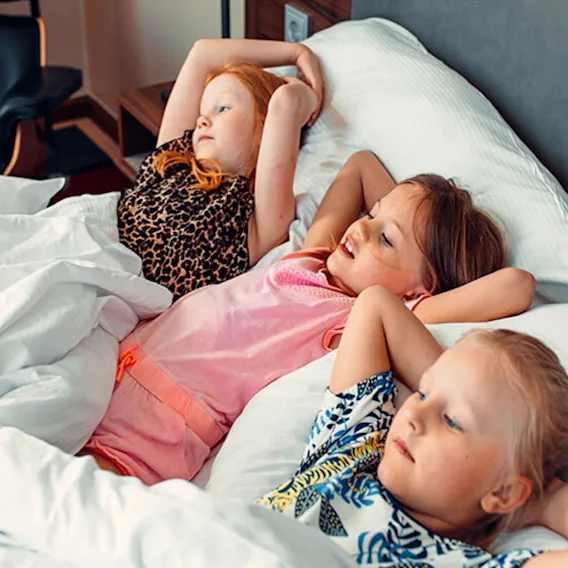 Tre barn kopplar av bekvämt i en säng med vita lakan, med händerna bakom huvudet, och njuter av en lugn stund.