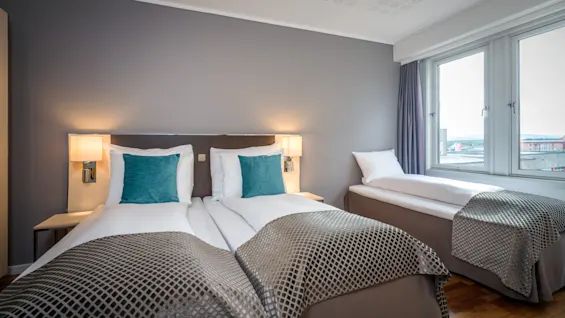Et komfortabelt hotellrom med tre senger, to sammen og en enkel, med turkise puter og et stort vindu.
