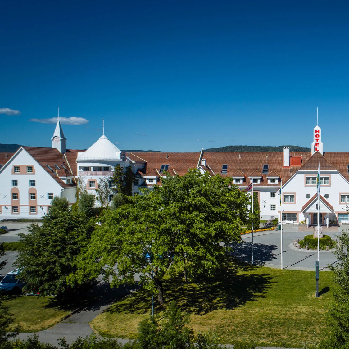 En stor hotelbygning med røde tegltage og en central indgang, omgivet af frodige træer og en klar blå himmel. Teksten lyder: "HOTEL" på et skilt over indgangen.